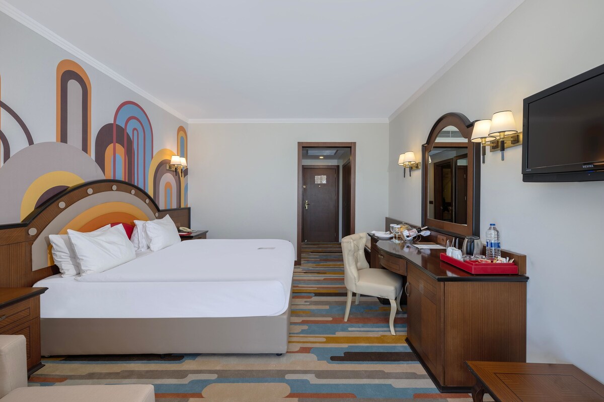 A hotel room with a large bed, a desk with a chair, a TV, and a bathroom door in the background.