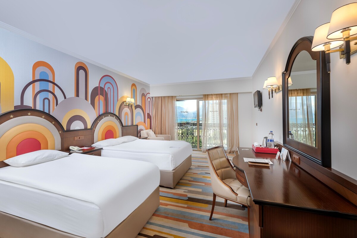A hotel room with two beds, a desk, a chair, a TV, and a balcony with a view.