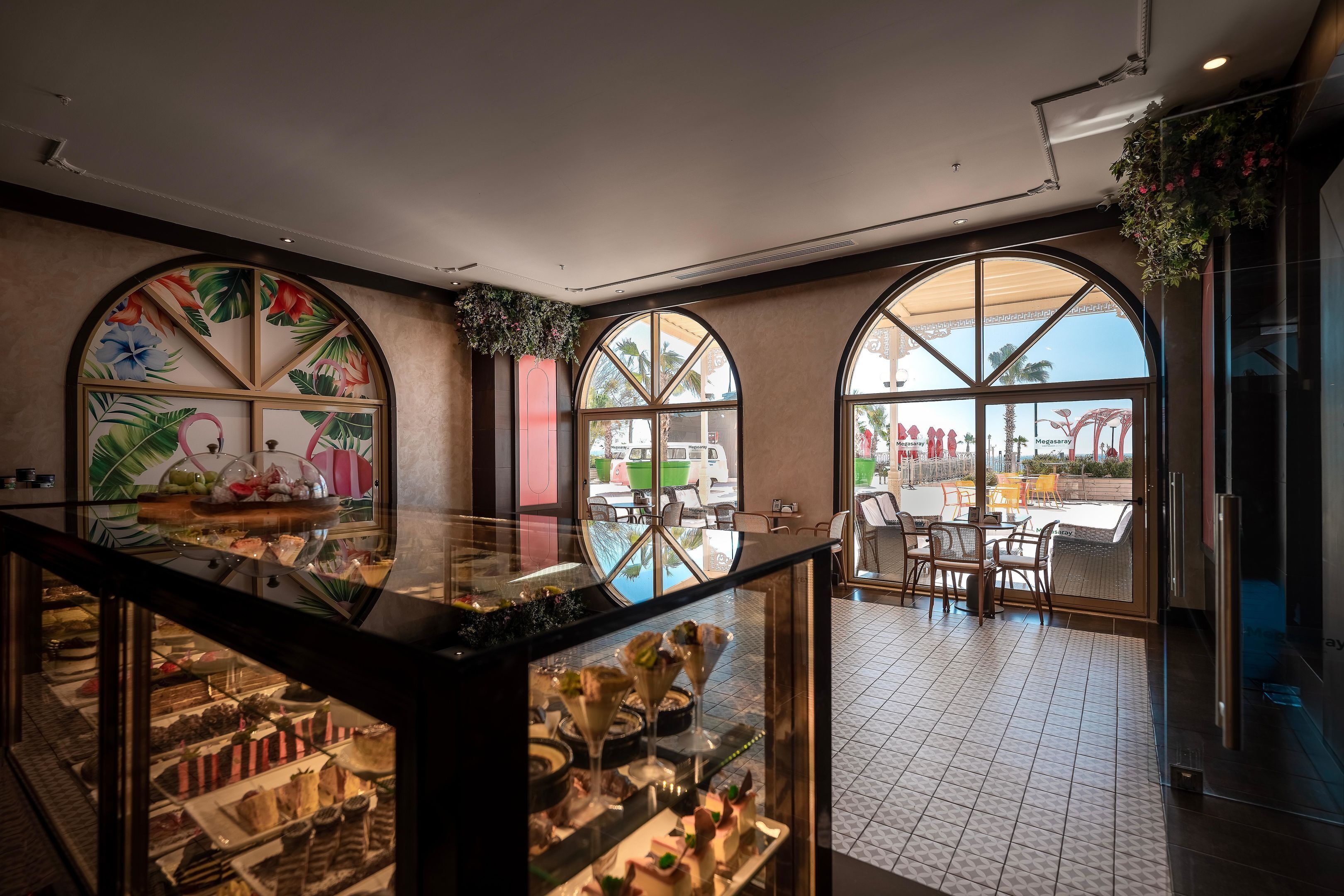Een hotel lobby met grote boogvensters, een glazen vitrine met desserts, en een zithoek met tafels en stoelen.