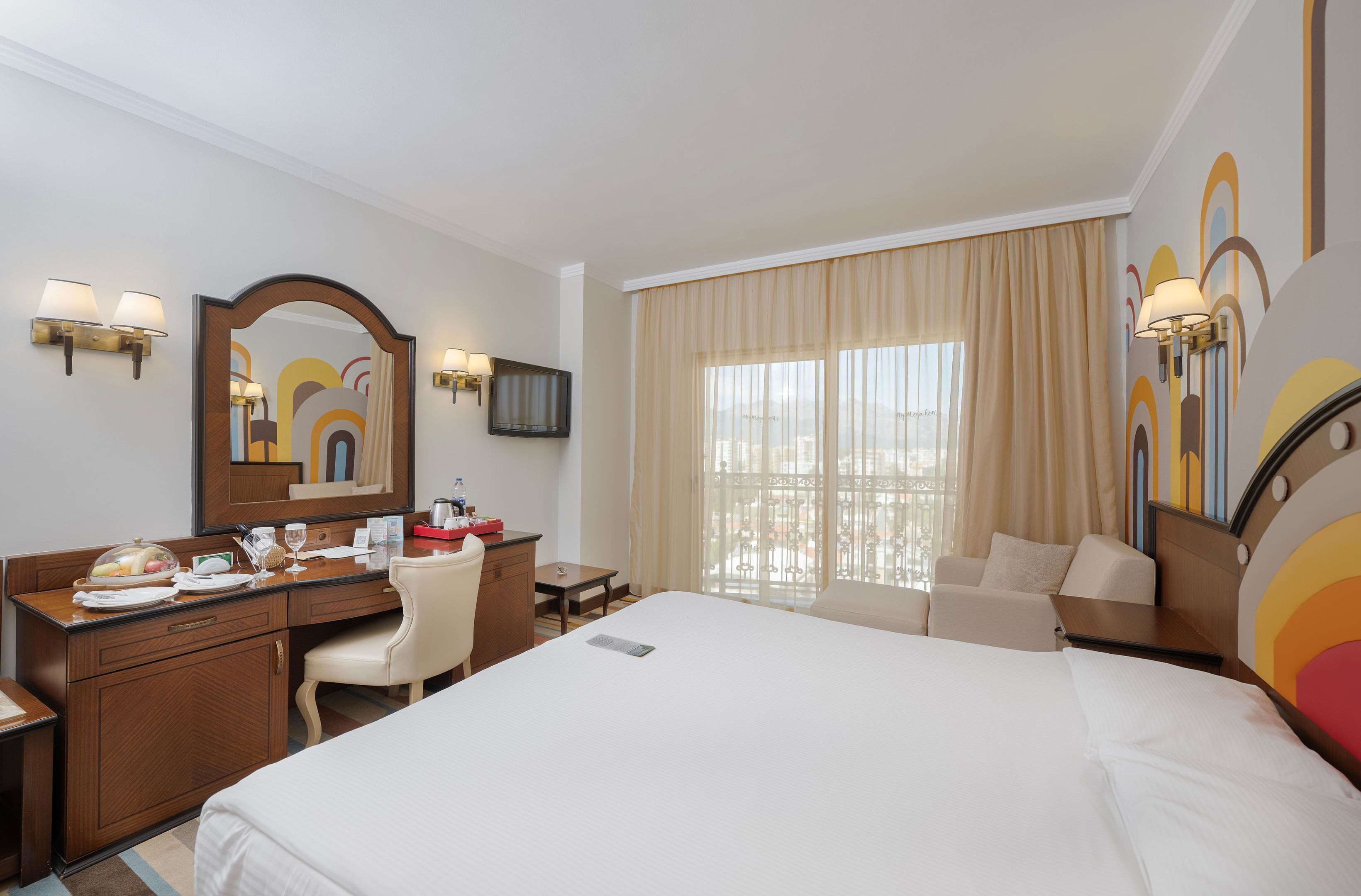 A hotel room with a large bed, a desk with a chair, a vanity with a mirror, and a window with curtains.