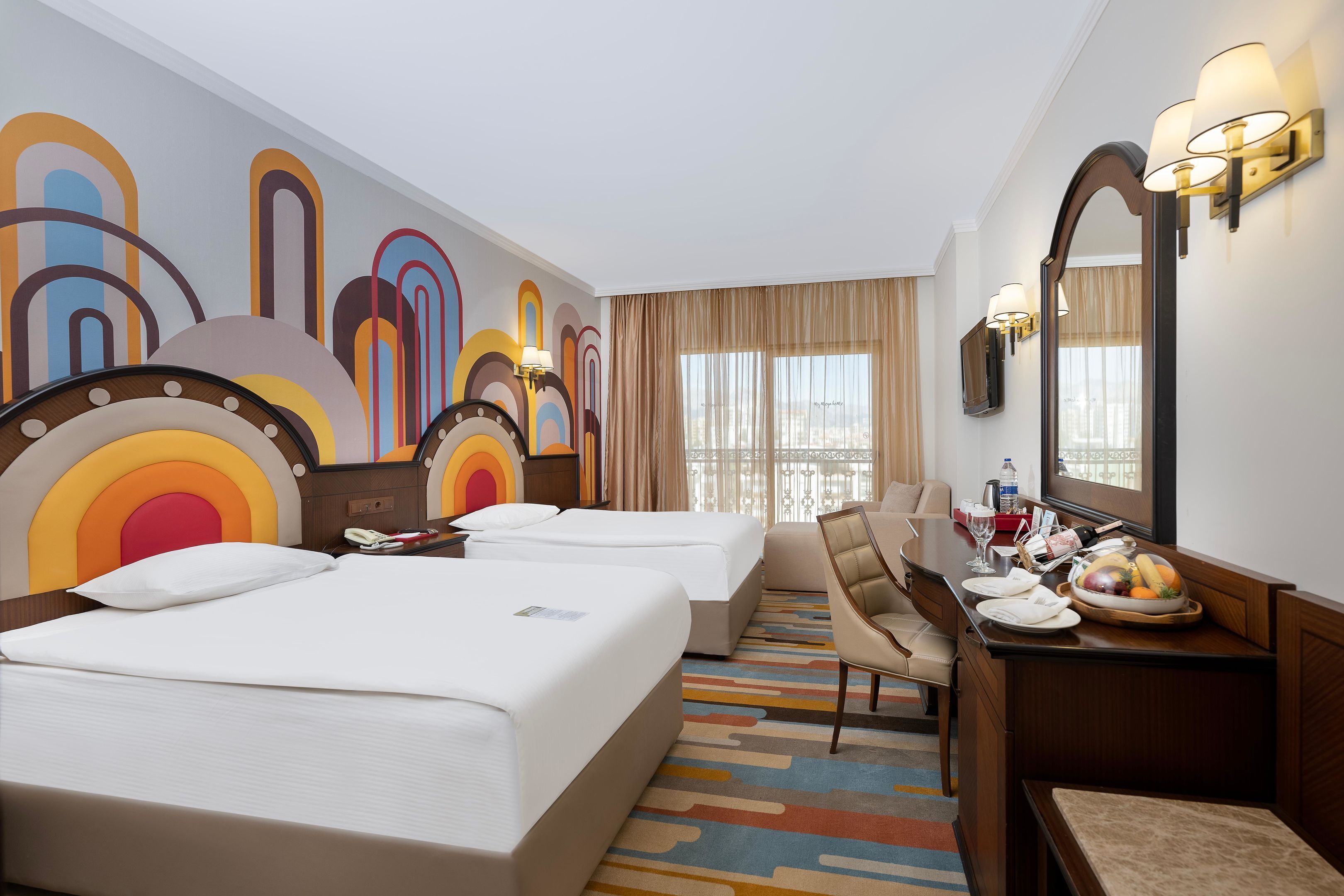 Habitación de hotel con dos camas, un escritorio con silla, un espejo y un mural abstracto de colores en la pared.