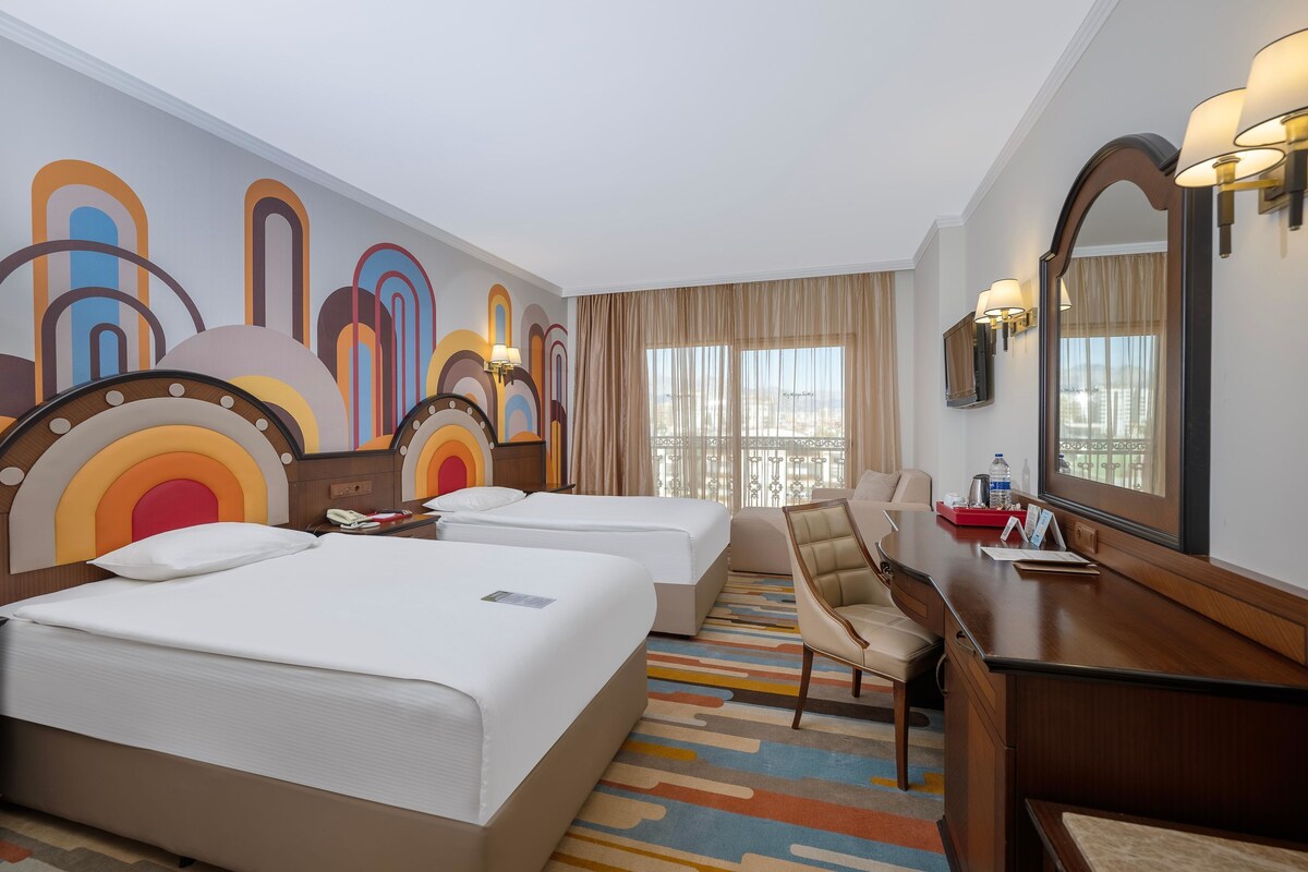 A hotel room with two beds, a desk, a chair, a large mirror, and a colorful abstract wall design.
