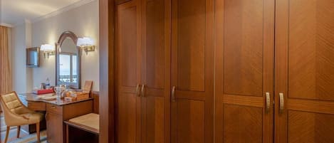 A hotel room with a large wooden wardrobe, a desk with a chair, a mirror with wall sconces, and a striped carpet.