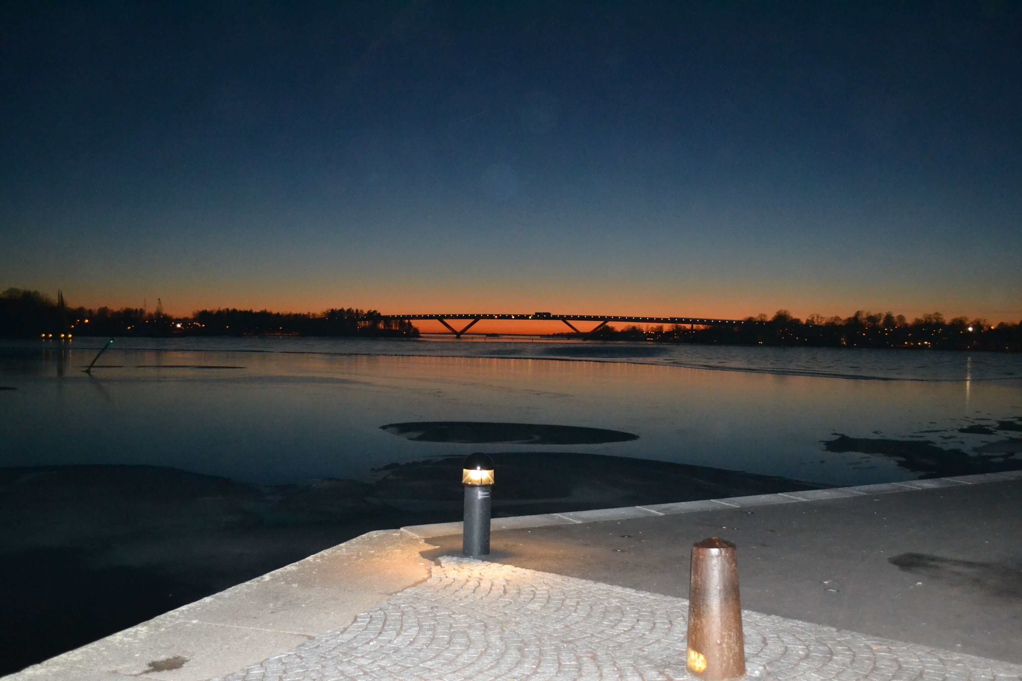 A nighttime view of a calm body of water with a bridge in the distance and a paved walkway in the foreground.