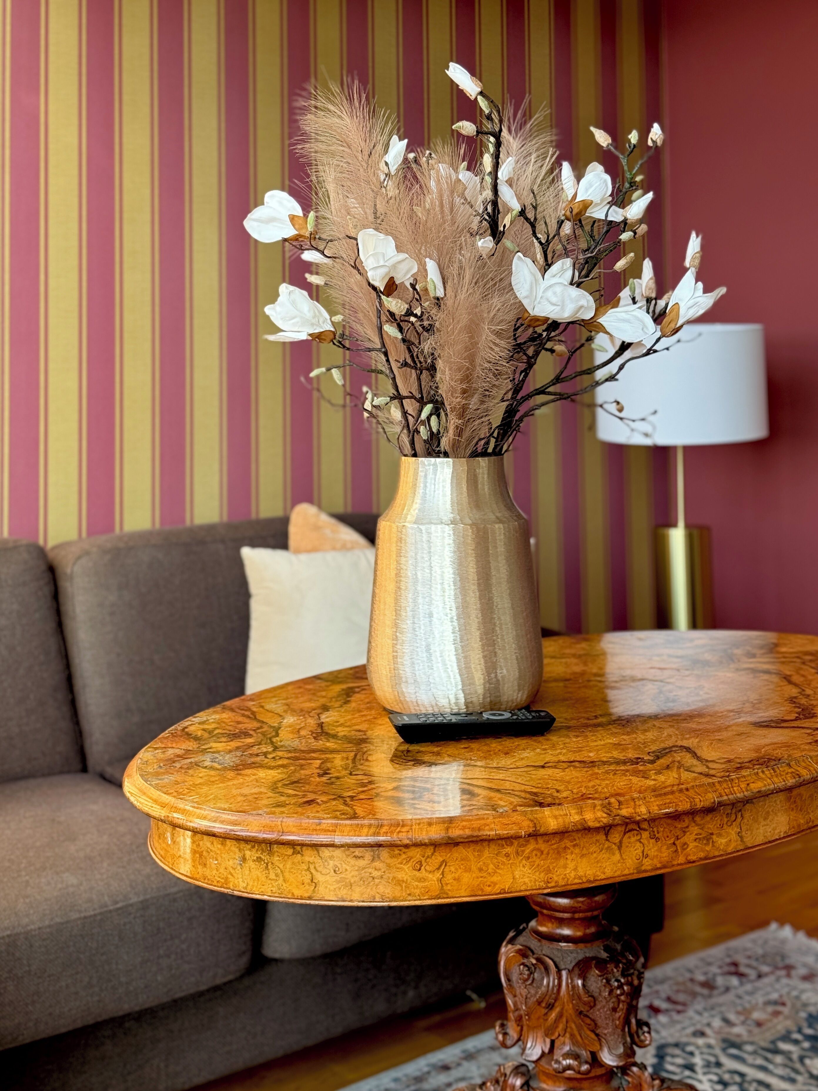 Een houten tafel met een vaas droog pampasgras en witte bloemen, een afstandsbediening en een lamp in een kamer met gestreept behang en een bank.