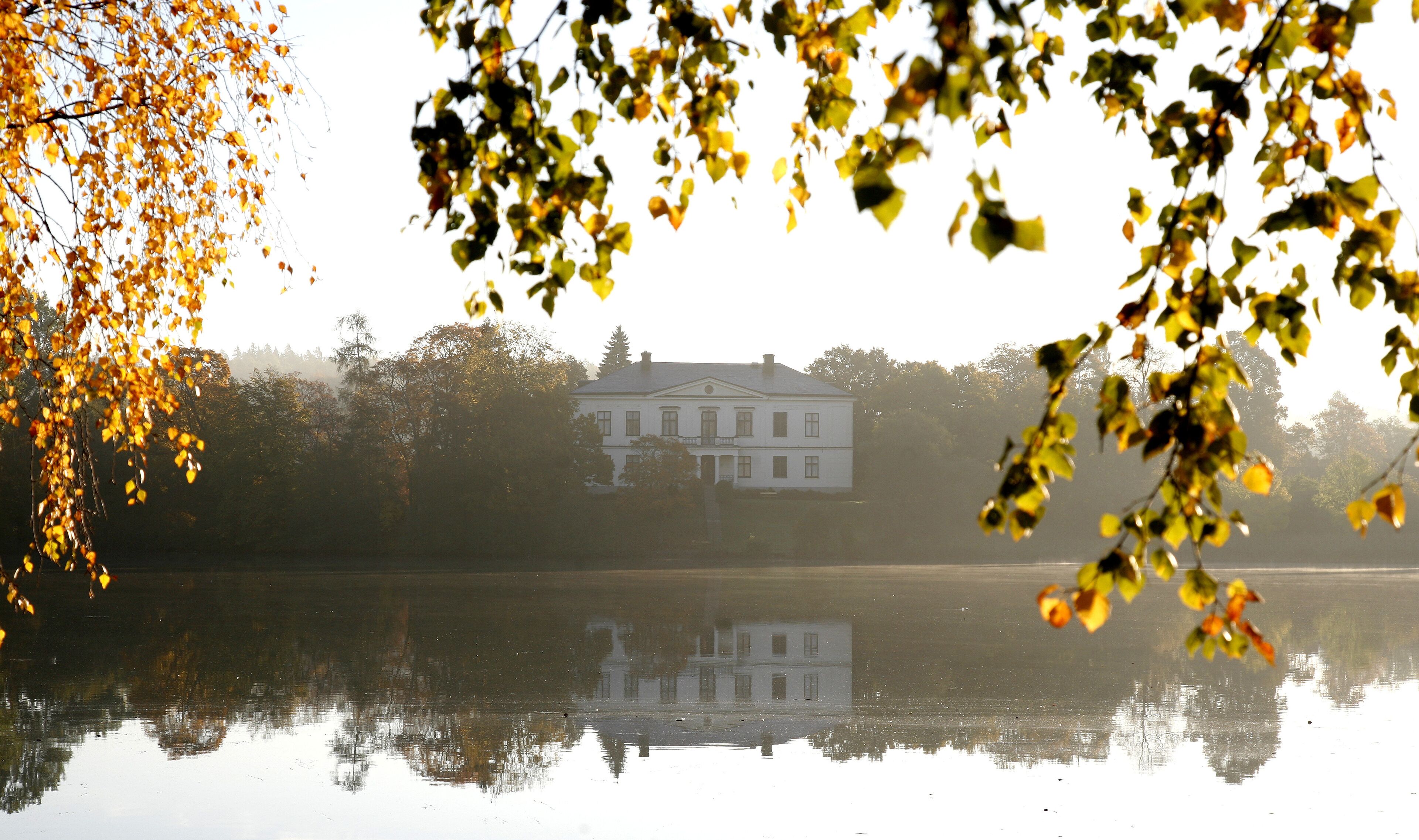A house reflected in a calm lake with autumn leaves in the foreground.