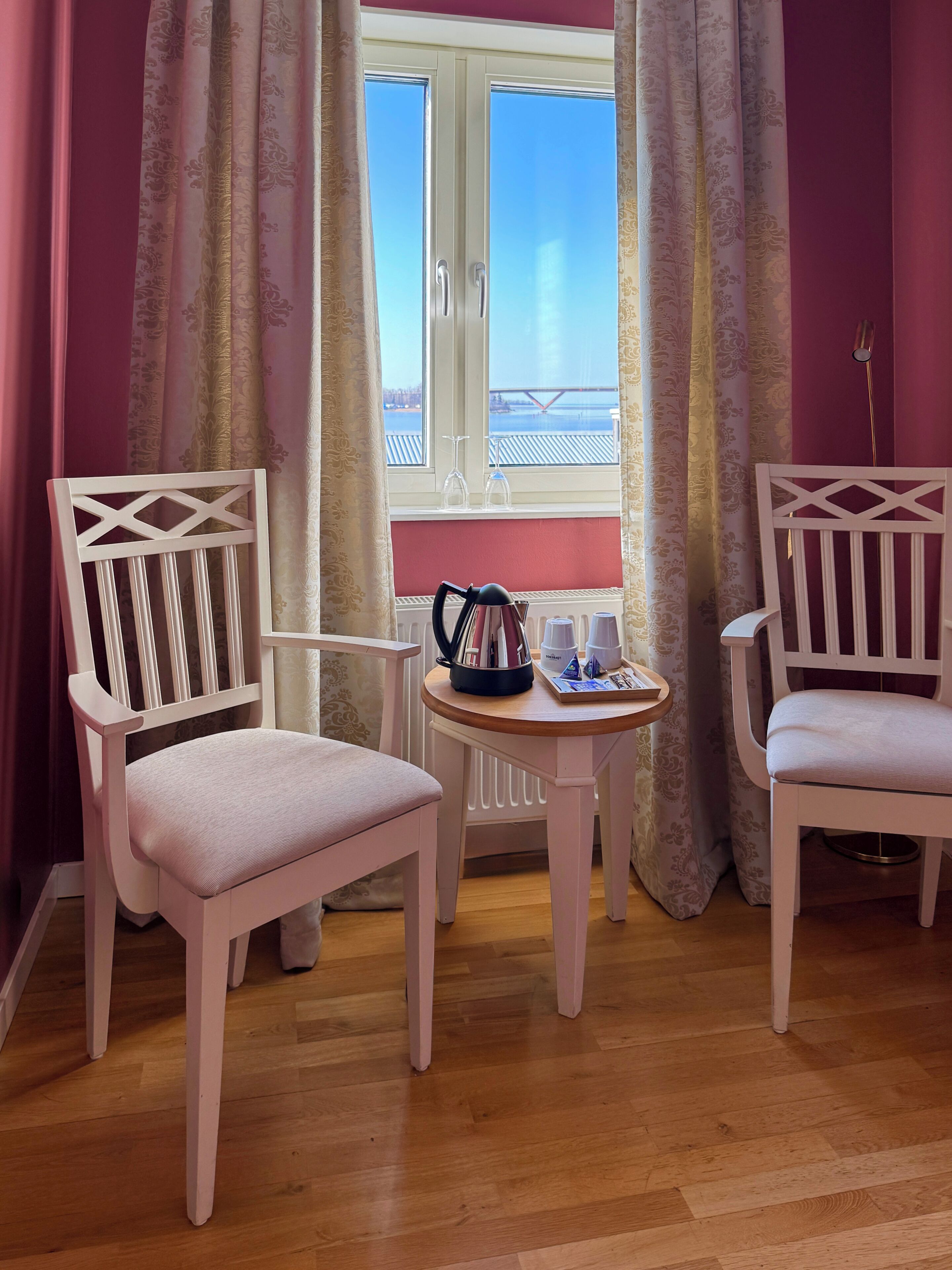 Dos sillas blancas con una mesa entre ellas, una ventana con cortinas y una vista de un puente.