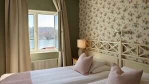 Habitación de hotel con dos camas, una ventana con cortinas, una lámpara y papel tapiz con motivos florales.