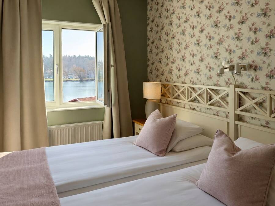 Quarto de hotel com duas camas, janela com cortinas, luminária e papel de parede floral.
