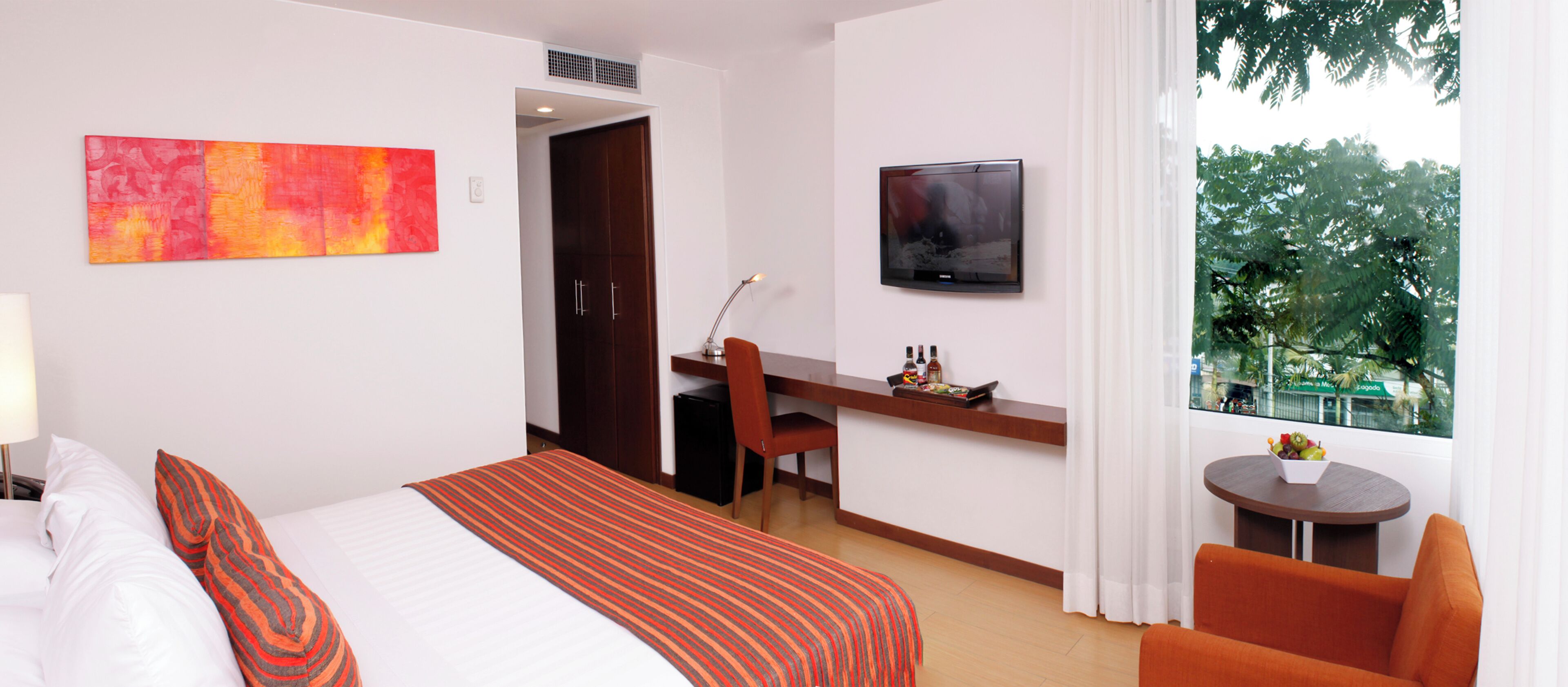 A hotel room with a bed, a desk, a chair, a TV, and a window with a view of trees.