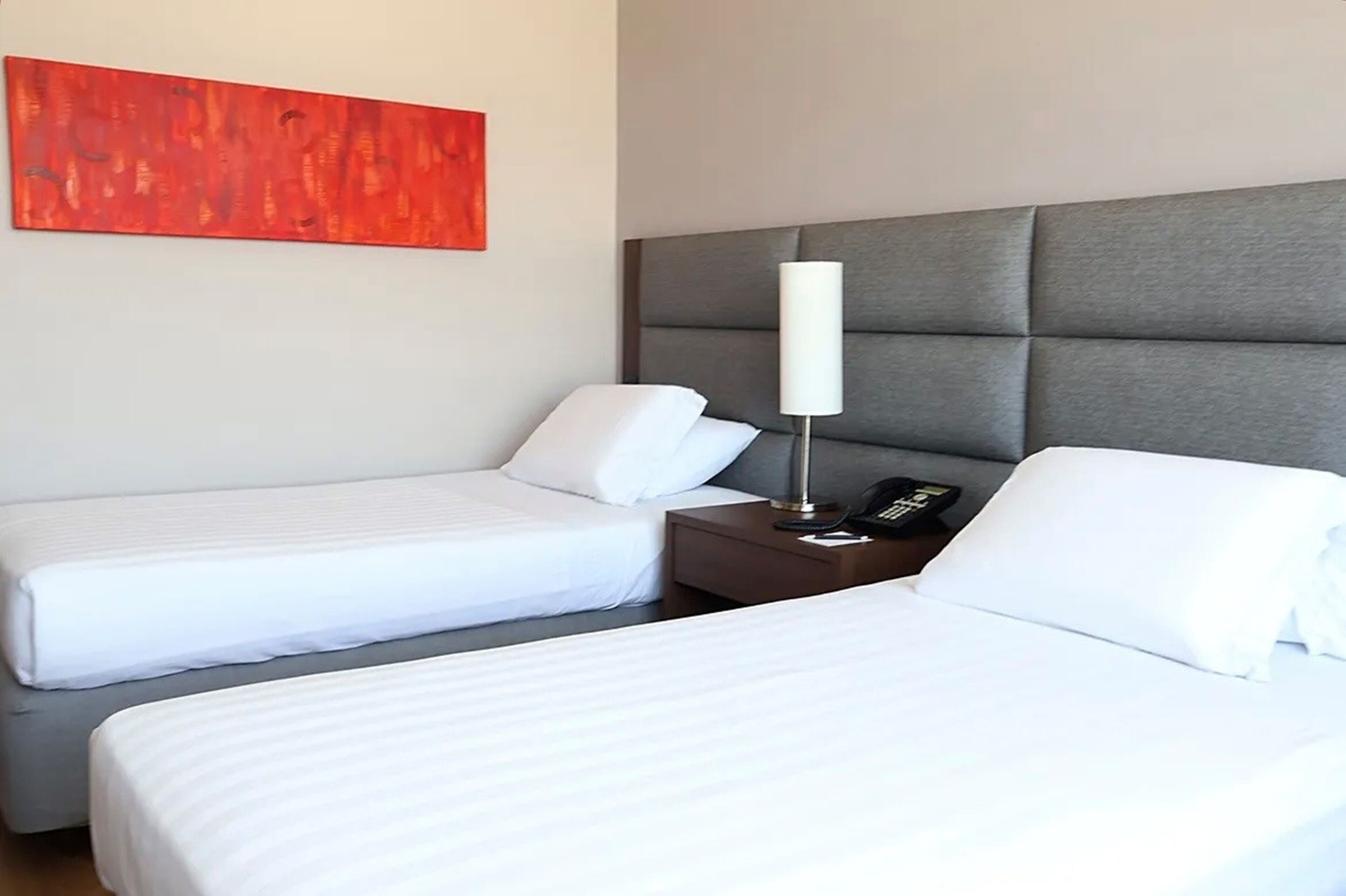 A hotel room with two beds, a grey headboard, a bedside table with a lamp, and a red abstract painting on the wall.
