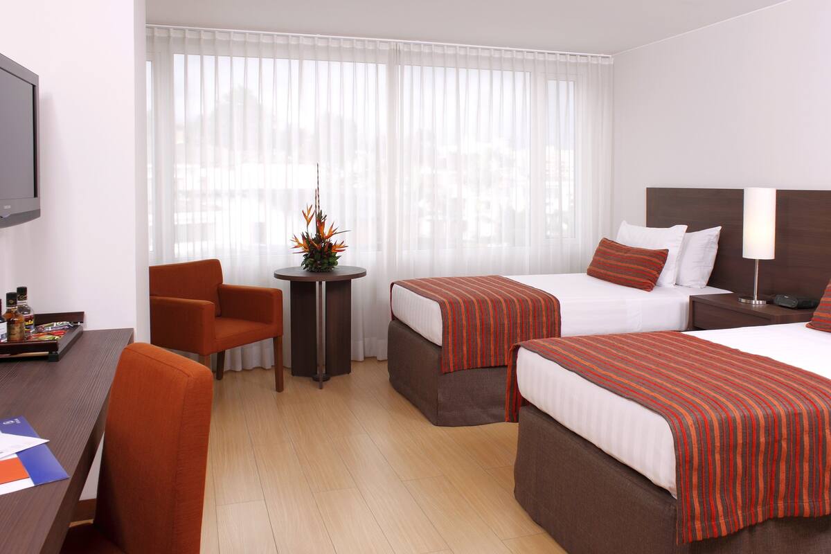 Ein Hotelzimmer mit zwei Betten, einem Schreibtisch mit Stuhl, einem kleinen Tisch mit Pflanze und einem Fernseher an der Wand.