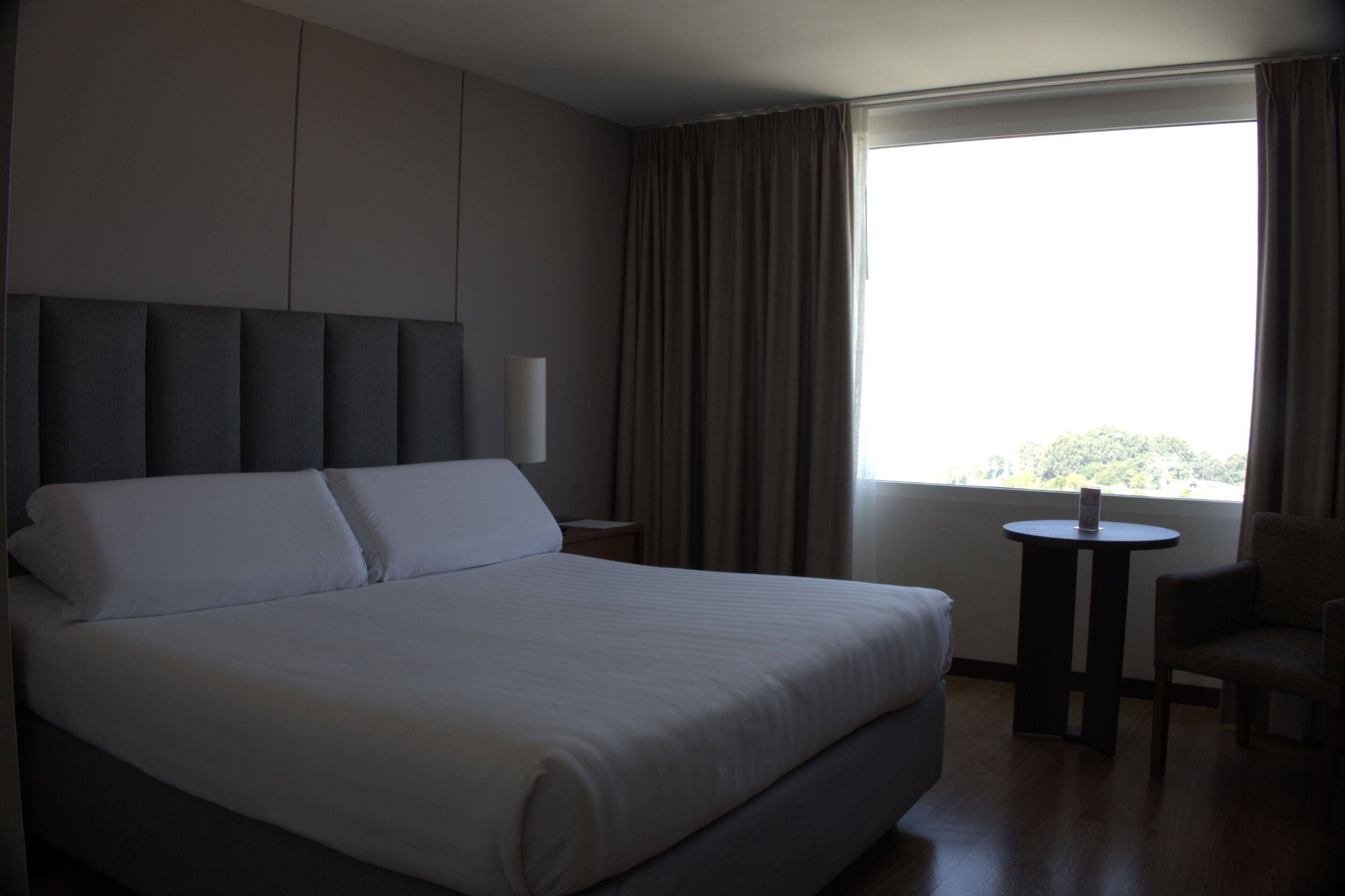 A hotel room with a large bed, a bedside table, a chair, and a window with curtains.