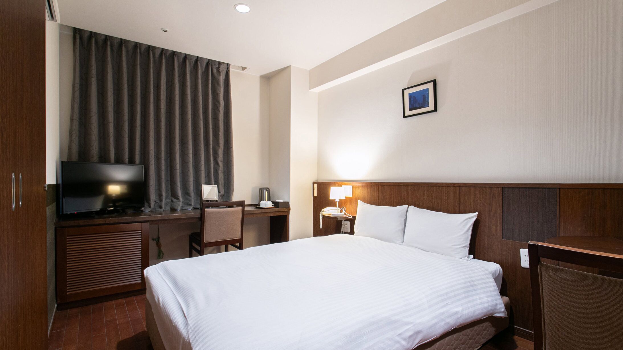 Quarto de hotel com uma cama, uma mesa com televisão, uma cadeira, um abajur e um quadro na parede.