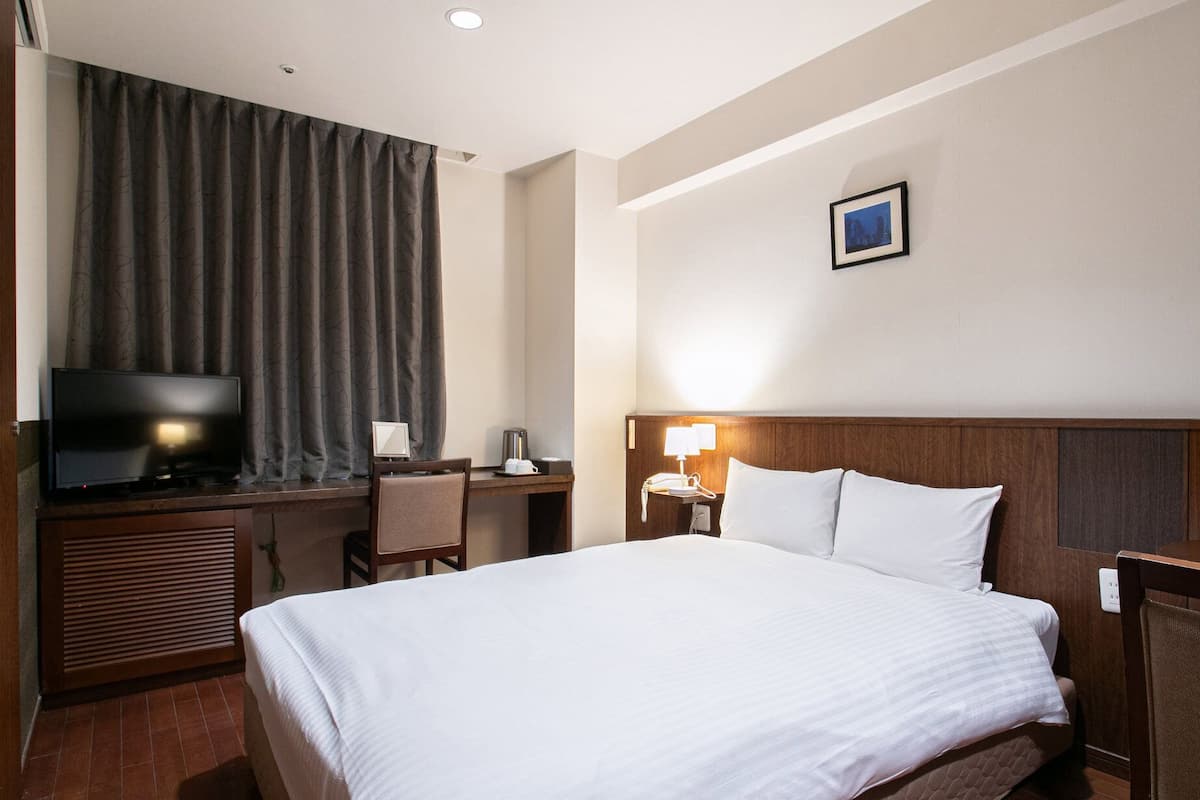 Quarto de hotel com uma cama, uma mesa com televisão, uma cadeira, um abajur e um quadro na parede.