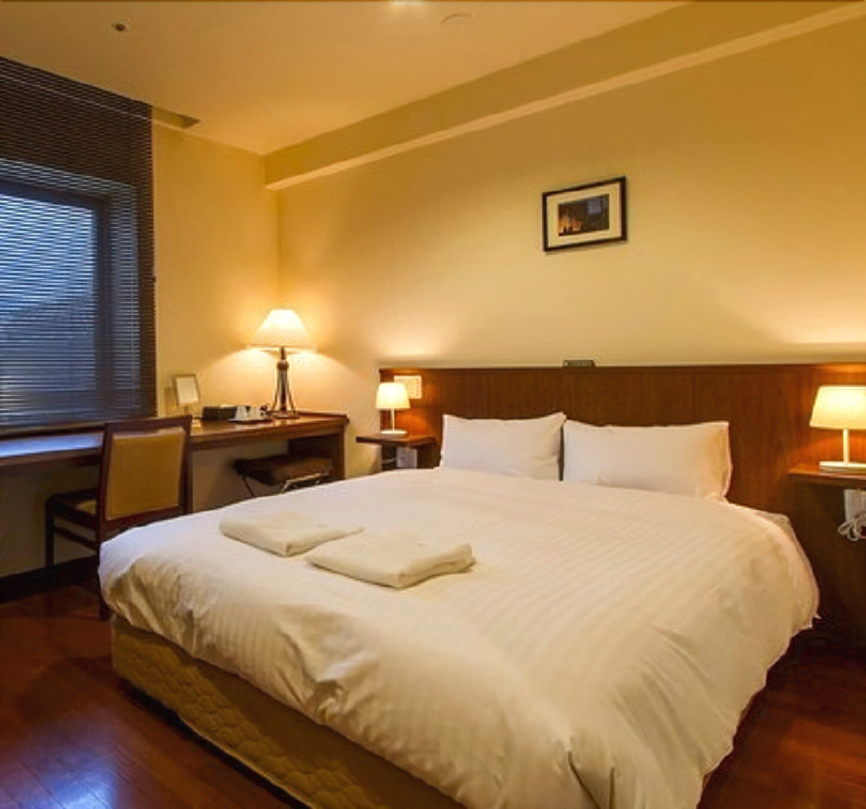 Ein Hotelzimmer mit einem großen Bett, zwei Nachttischlampen, einem Schreibtisch mit Stuhl und einem gerahmten Bild an der Wand.