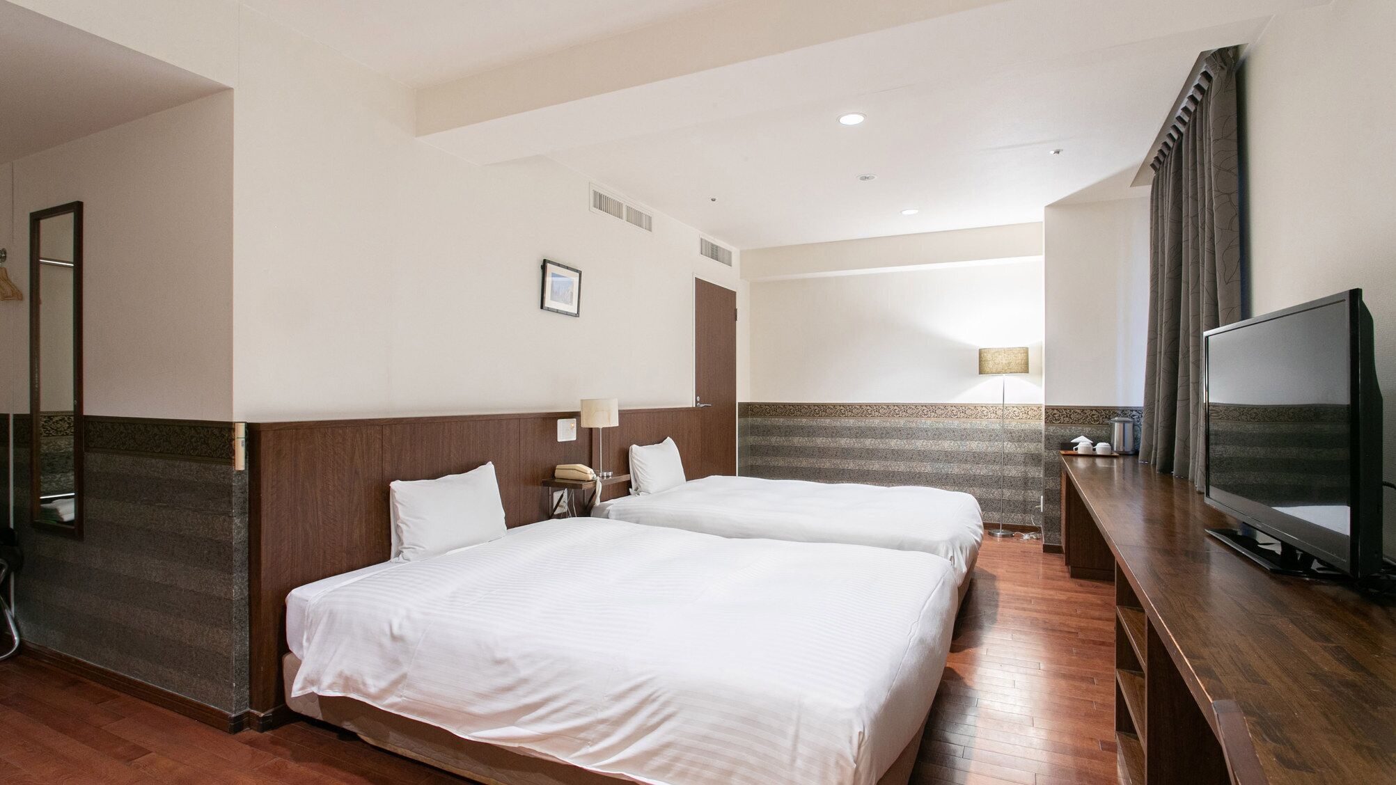Quarto de hotel com duas camas, uma televisão, uma escrivaninha e banheiro com espelho.