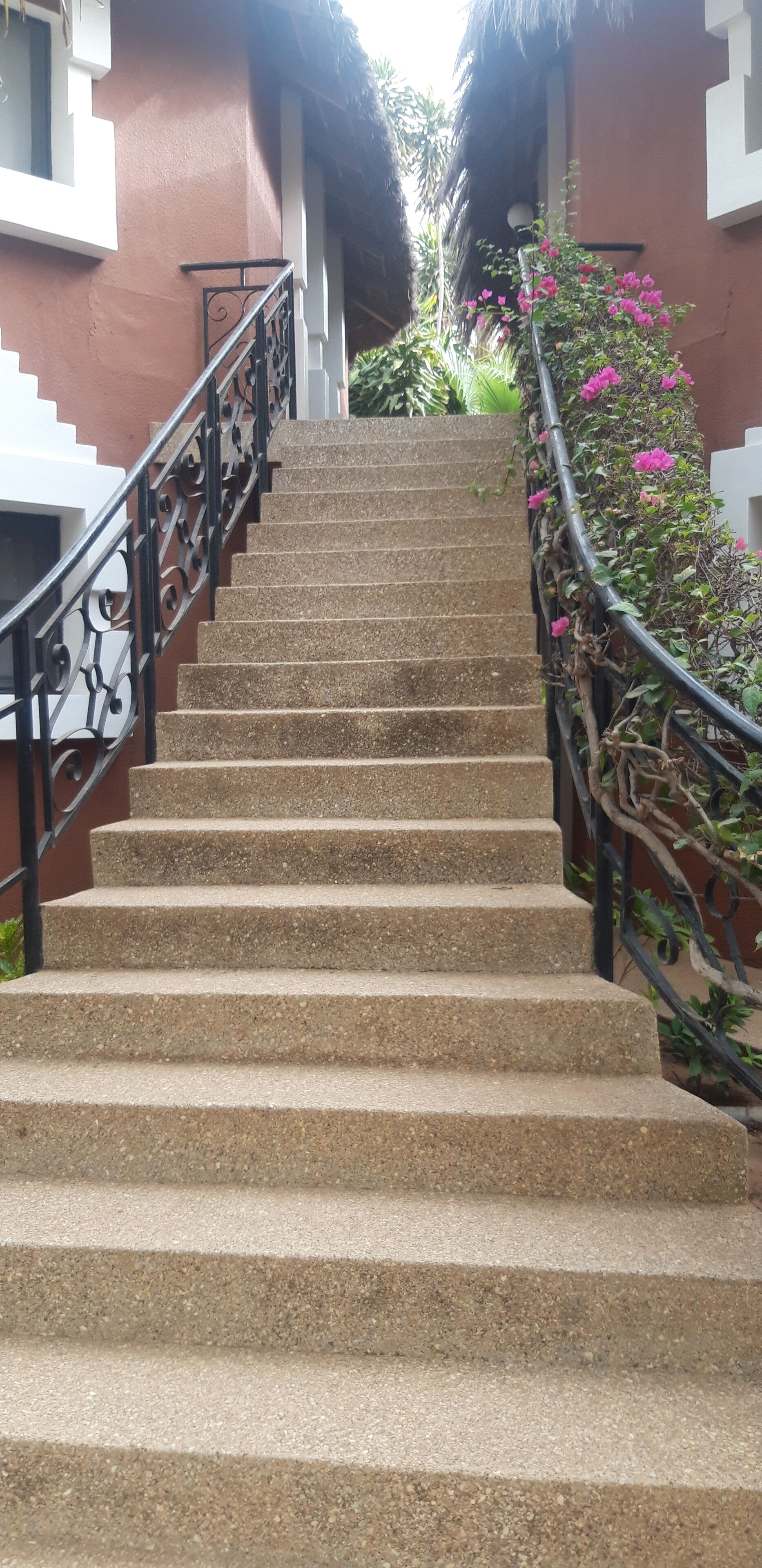 A staircase with stone steps and a metal railing, leading to a building with a red exterior and a thatched roof.