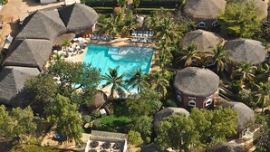 Vue aérienne d’un complexe hôtelier doté d’une piscine centrale, de bungalows aux toits de chaume et de palmiers.