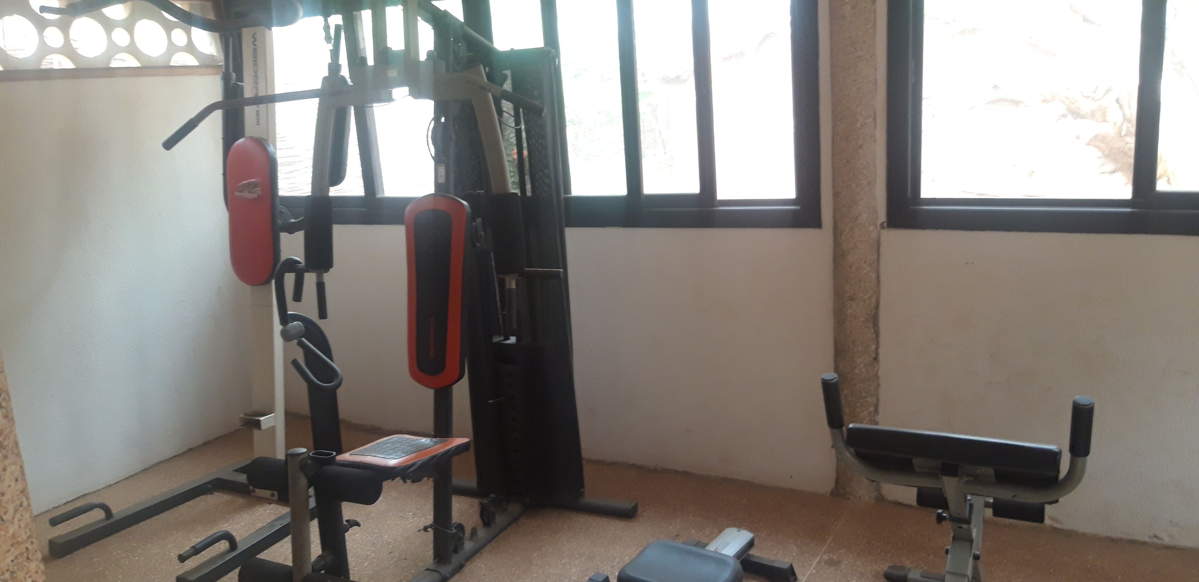 Une salle de sport équipée de matériel d’exercice, d’un banc et d’une fenêtre avec des stores.