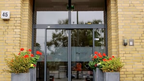 L’extérieur de l’hôtel H+Hotel Youth Berlin se distingue par sa façade en briques, son grand hall d’entrée vitré et ses deux jardinières ornées de fleurs rouges.