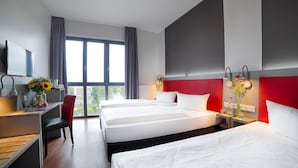Une chambre d’hôtel avec trois lits, un bureau avec un ordinateur, une chaise, une télévision et de grandes fenêtres avec des rideaux.