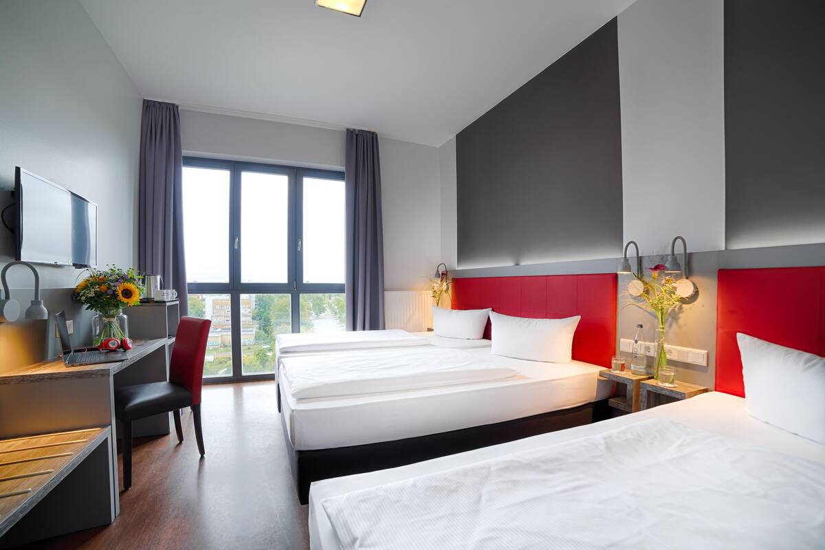 Hotelkamer met drie bedden, een bureau met een computer, een stoel, een televisie en grote ramen met gordijnen.
