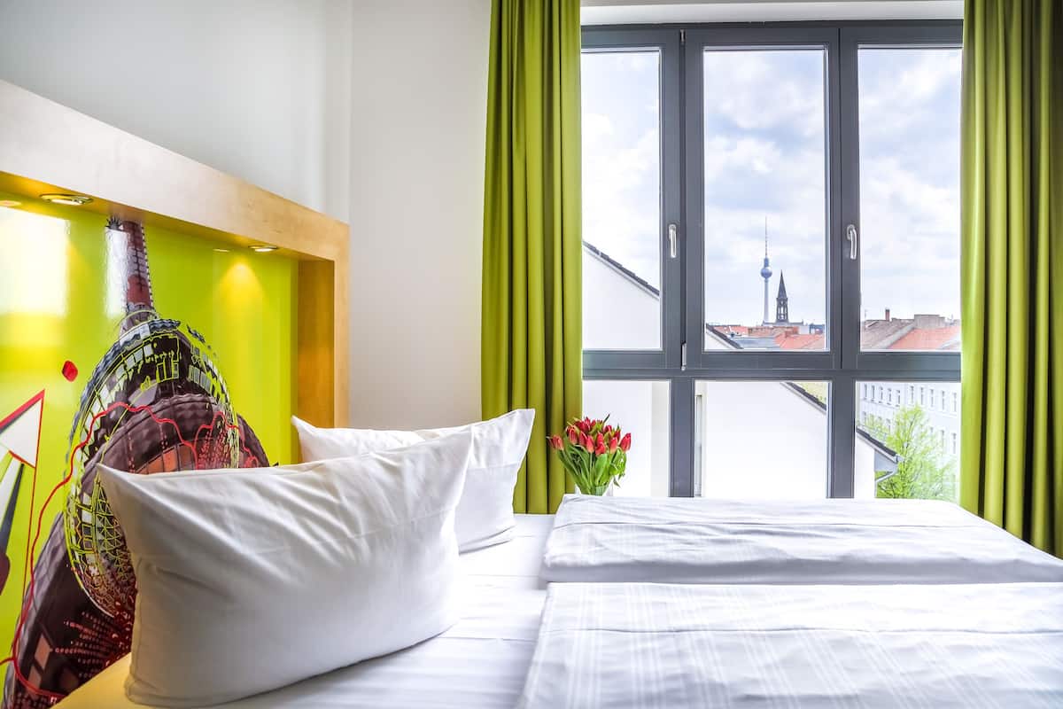 Hotelkamer met een bed, groene gordijnen, een raam met uitzicht op de stad en een decoratief wandobject.
