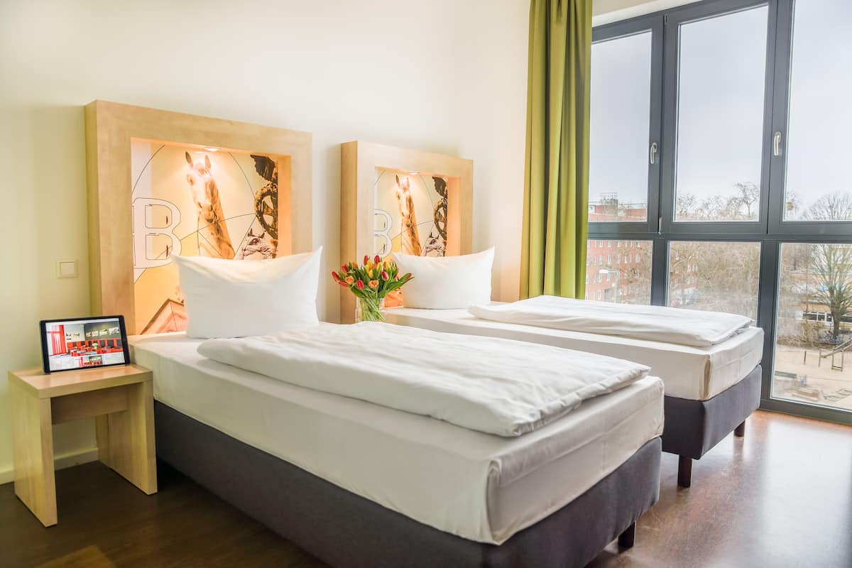 Une chambre d’hôtel avec deux lits, une table de chevet équipée d’une tablette et une grande fenêtre donnant sur des immeubles.