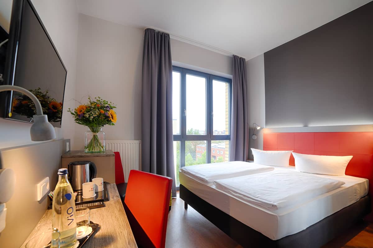 Een hotelkamer met een groot bed, een bureau met stoel, een spiegel, een raam met gordijnen en een vaas met bloemen.