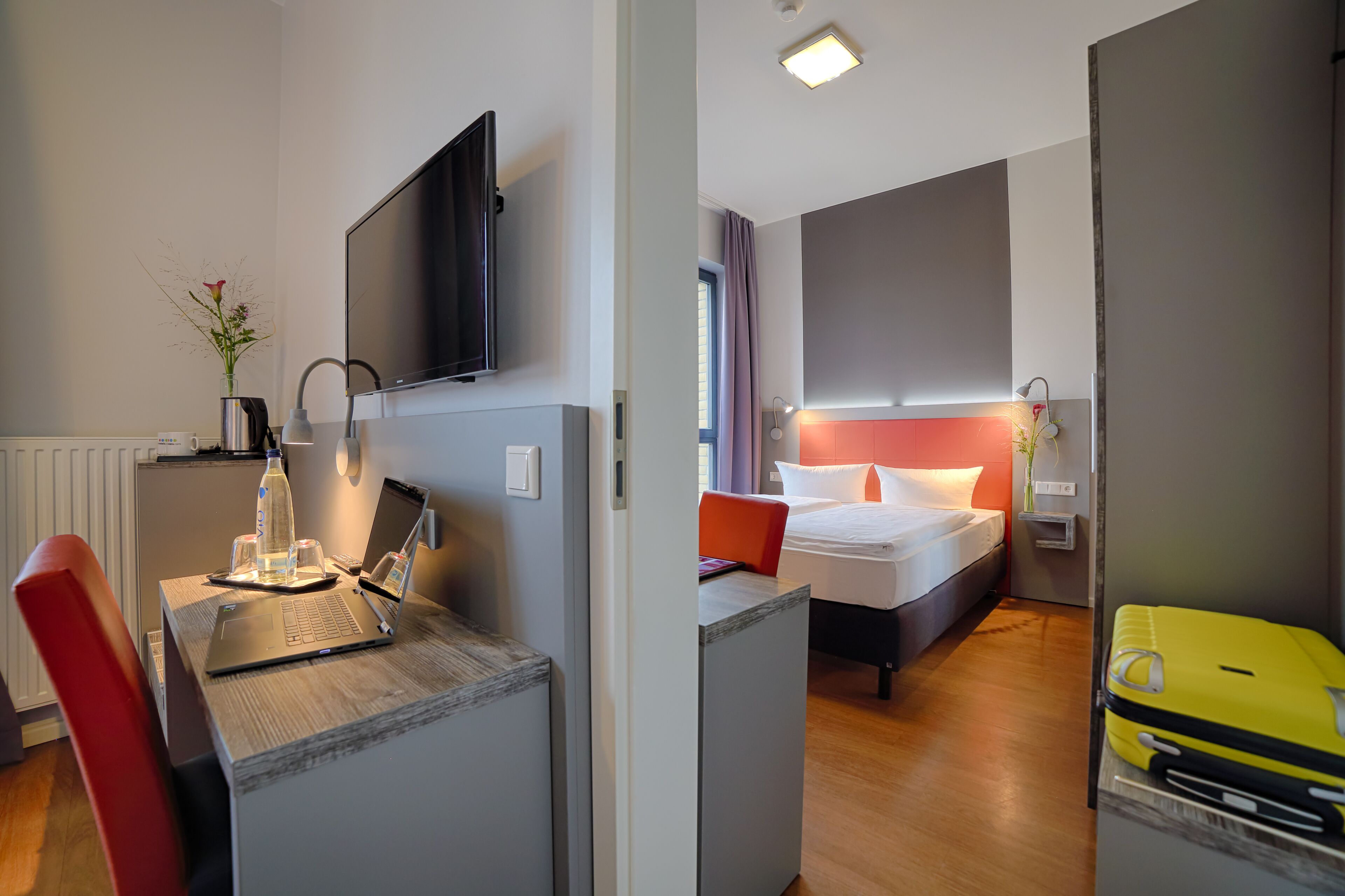 A hotel room with a desk, laptop, and a bed with a red headboard.