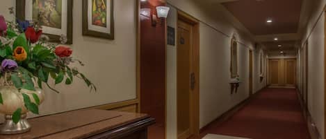 A hotel hallway with a marble reception desk, framed artwork on the walls, a potted plant, and a red carpeted floor.
