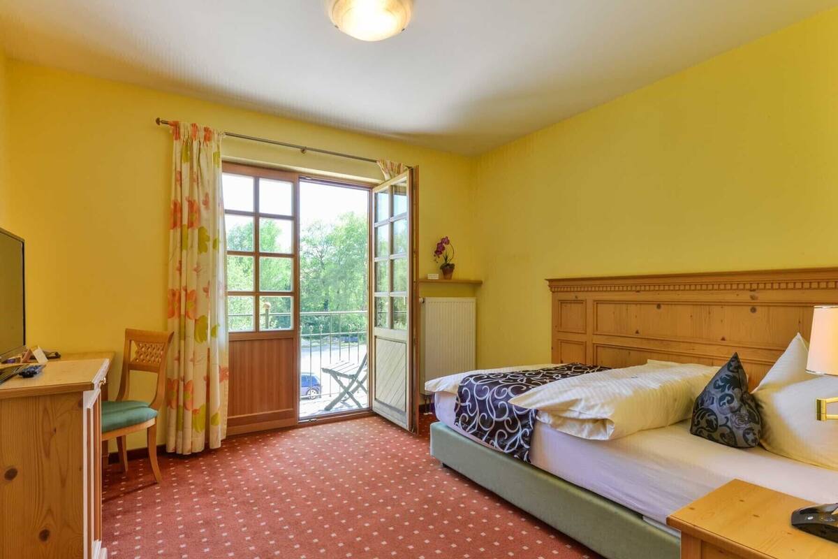 Een slaapkamer met een bed, een bureau, een stoel, een raam met gordijnen en uitzicht op groen.