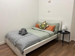 A single bed with a grey blanket and pillows, a bedside table with a lamp, and a white wall with a power outlet.
