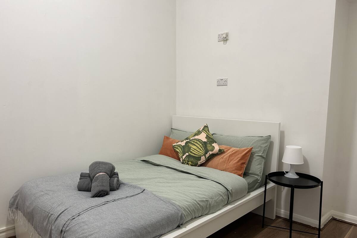 A single bed with a grey blanket, a green and orange pillow, and a lamp on a small black table beside it.