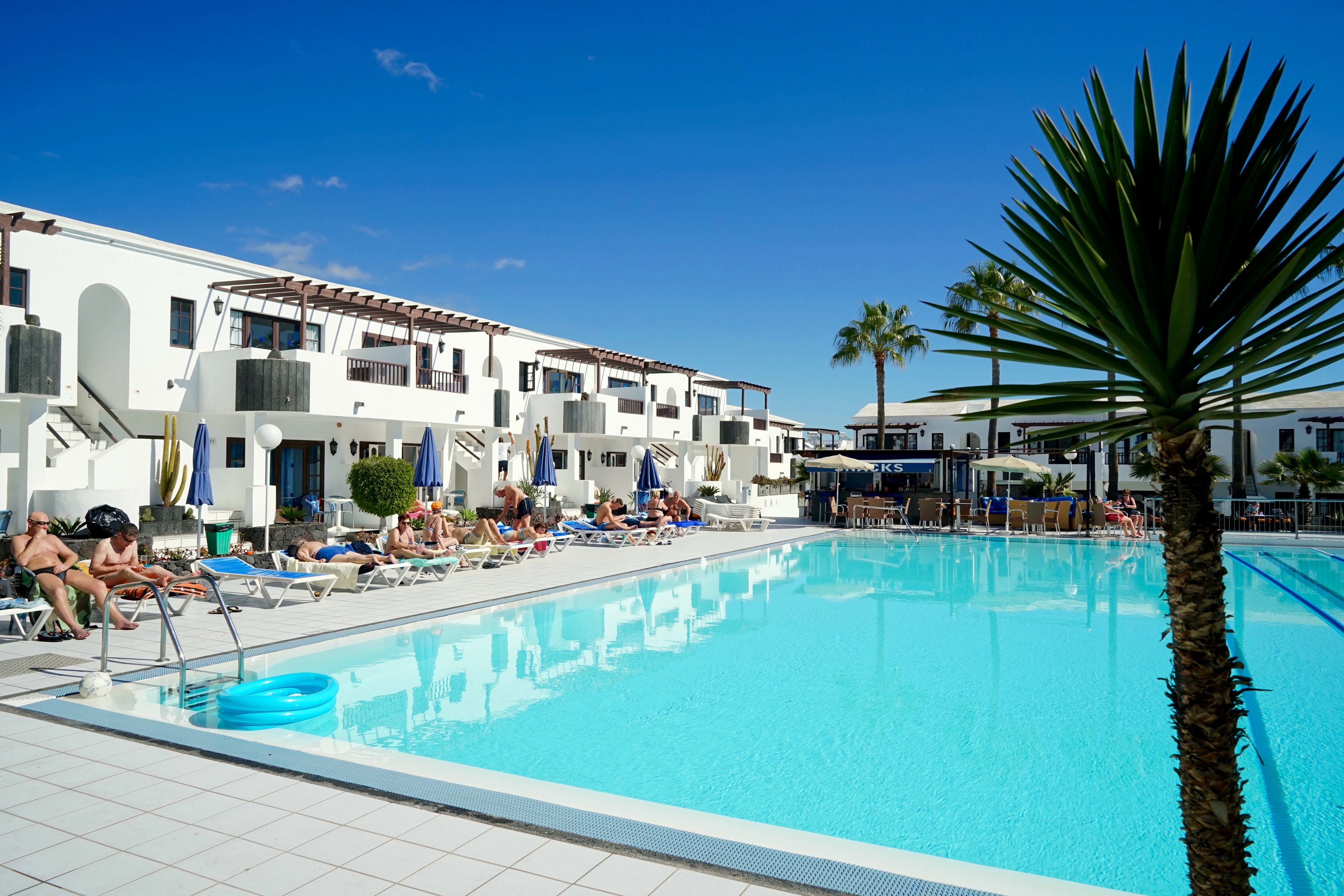 Piscina com espreguiçadeiras e guarda-sóis, rodeada por edifícios brancos com varandas.