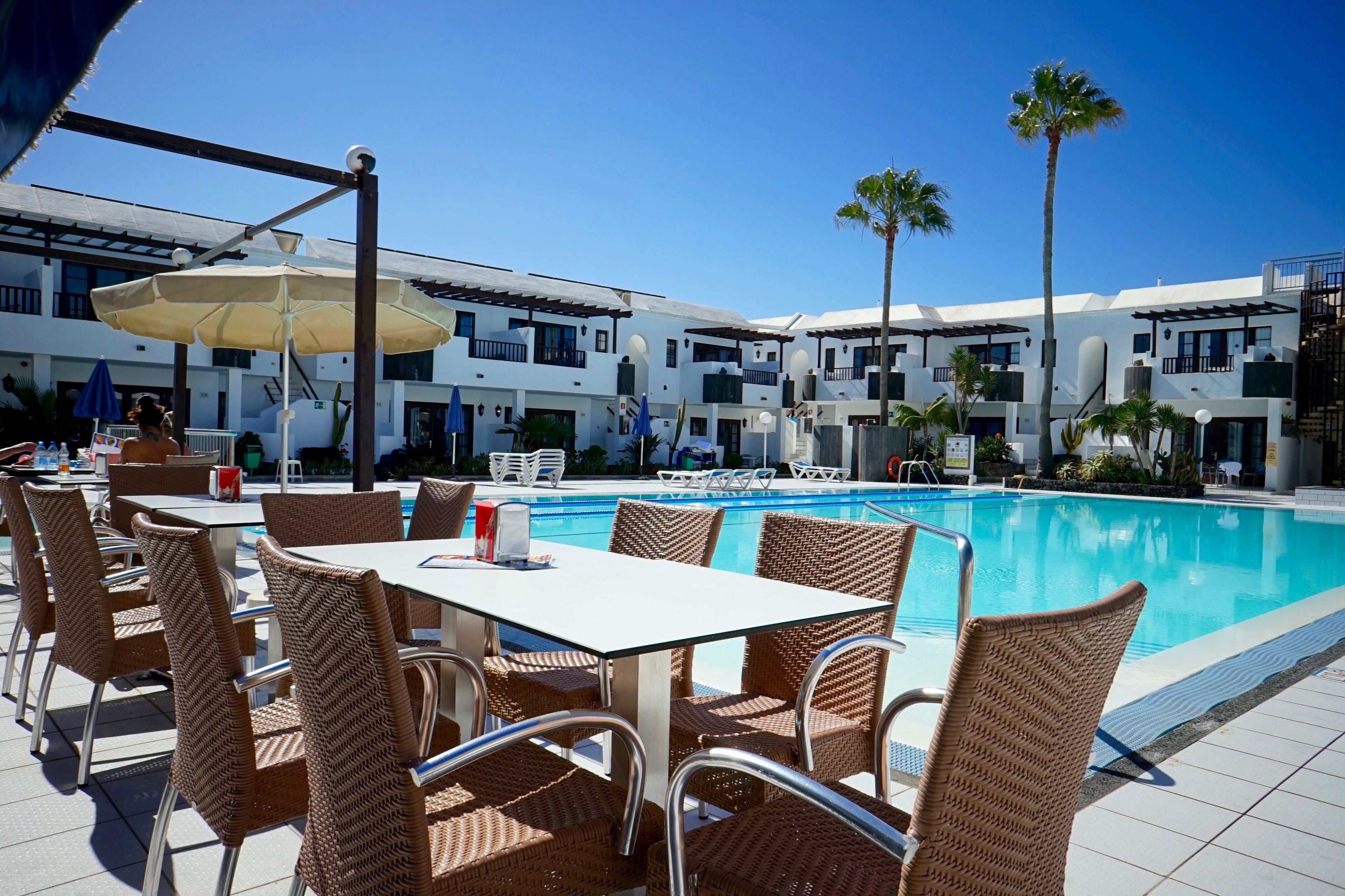 A poolside area with wicker chairs and tables, a pool, and a building with balconies.