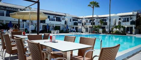 A poolside area with wicker chairs and tables, a pool, and a building with balconies.