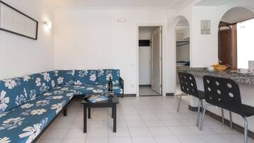 Een woonkamer met een blauwe bloemenbank, een tafeltje met een fles en glazen, en een keuken met een aanrecht en stoelen.