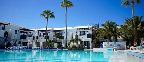 A swimming pool with lounge chairs and palm trees, surrounded by white buildings with balconies.