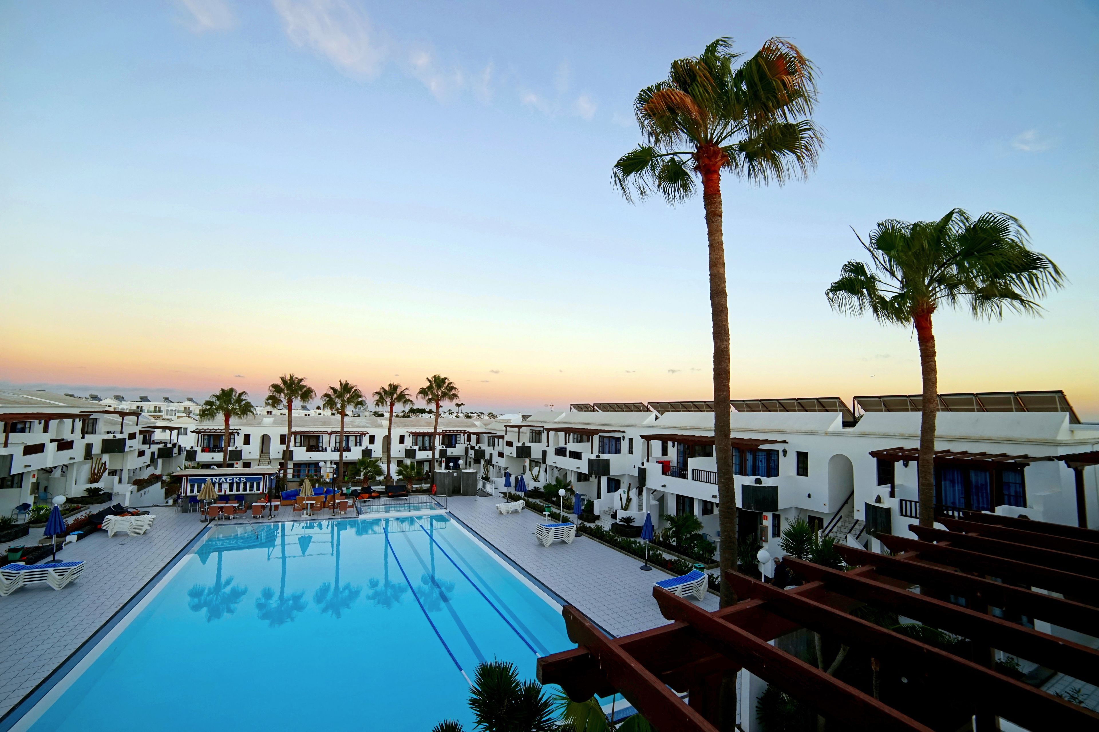 A resort pool area with multiple lounge chairs and umbrellas, surrounded by white buildings with balconies and palm trees.