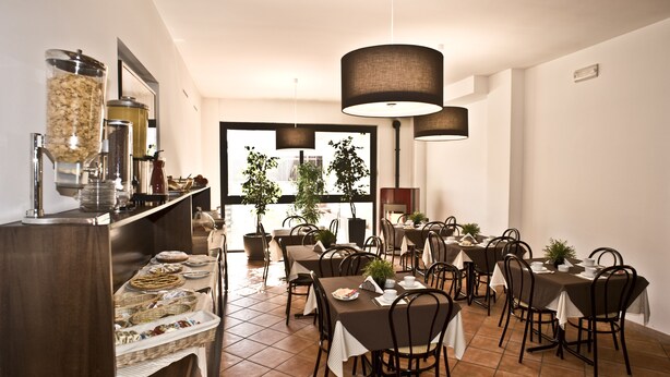 A dining area with multiple tables and chairs, a buffet with various food items, and large windows with a view of buildings outside. - Villa Santa Croce (San Giovanni Rotondo)