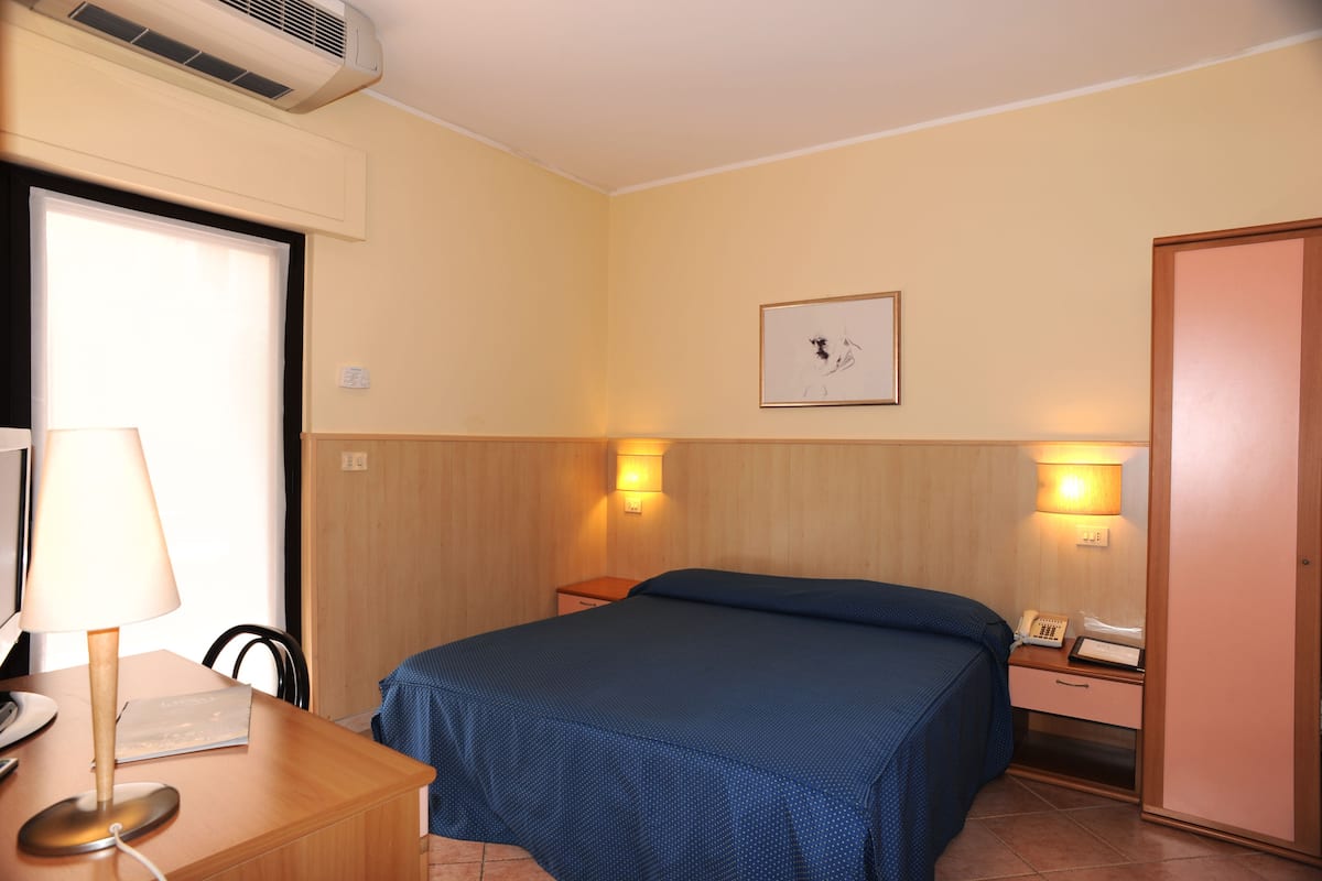 A hotel room with a bed, bedside tables, a desk with a lamp, a phone, and a wall-mounted picture.