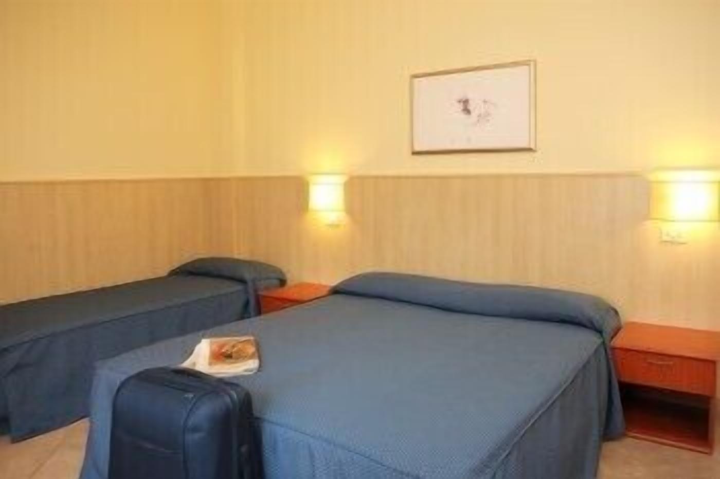 A hotel room with two single beds, a small table, a chair, a suitcase, and a framed picture on the wall.