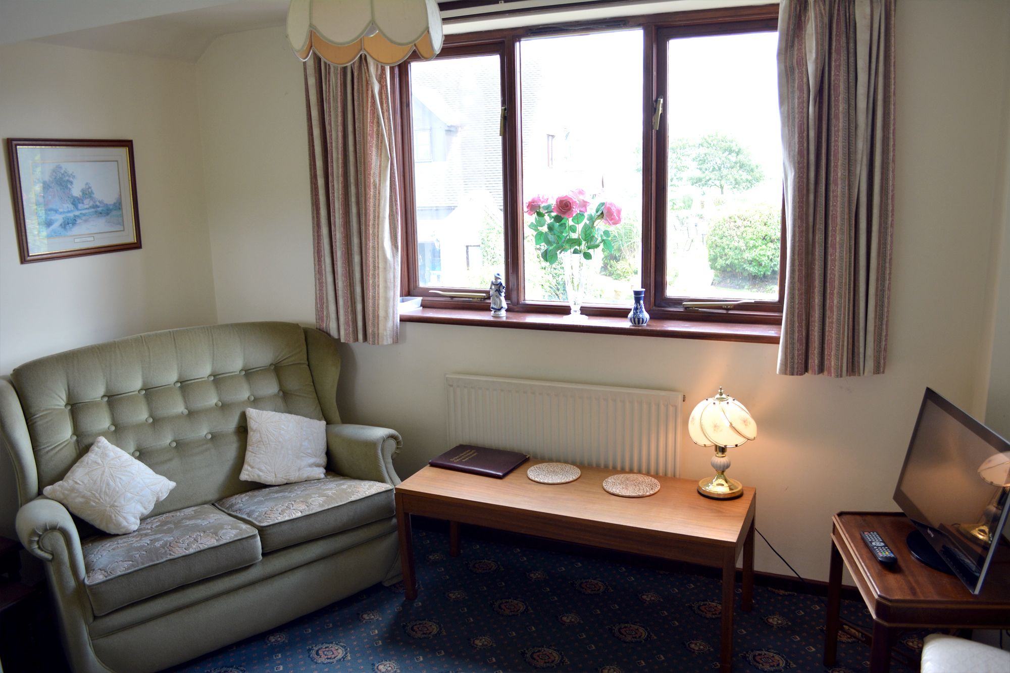A living room with a green sofa, a small table with a lamp, and a window with curtains.