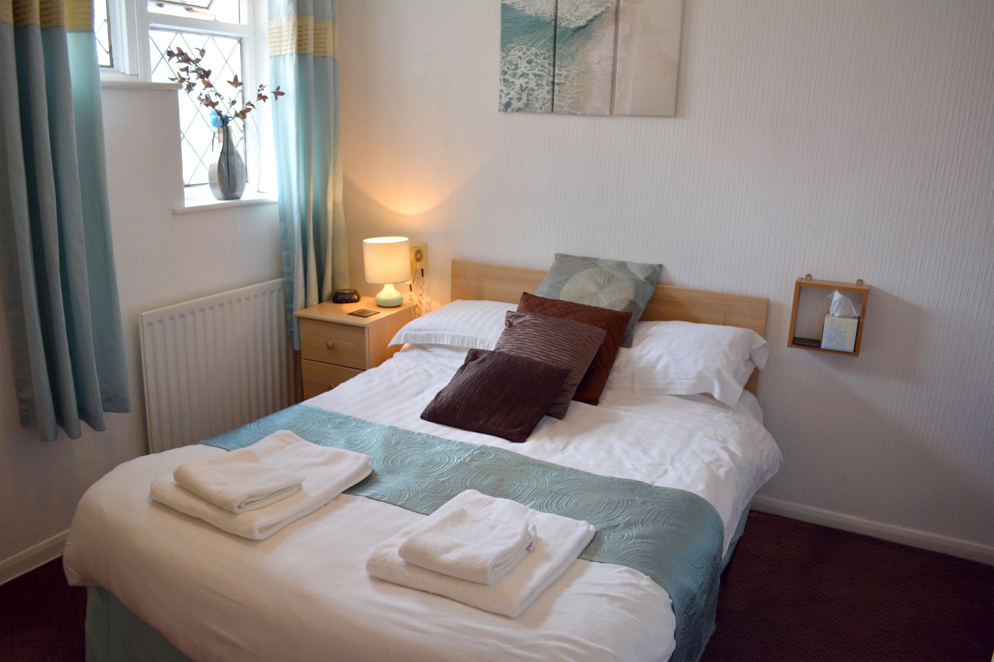 Een netjes opgemaakt bed met witte lakens en een blauwgroene sprei, een nachtkastje met een lamp, en een raam met gordijnen.