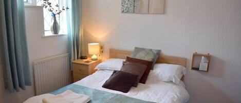 Een netjes opgemaakt bed met witte lakens en een blauwgroene sprei, een nachtkastje met een lamp, en een raam met gordijnen.