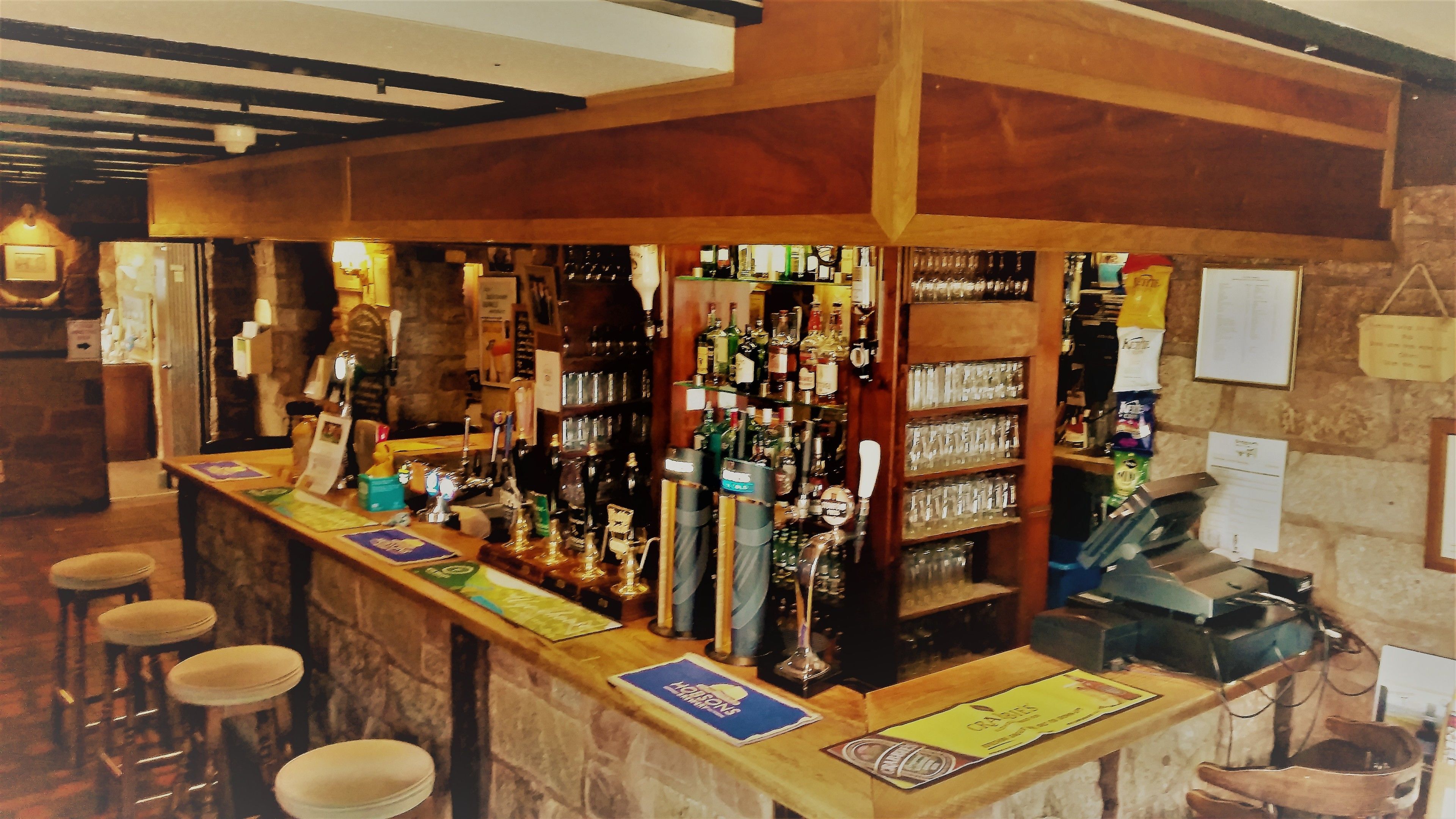 A bar with a wooden counter, stools, and shelves stocked with bottles and glasses.