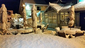 A snow-covered outdoor seating area with tables and chairs, a stone building in the background, and a glass door leading to an interior space. - The Bull's Head Inn at Chelmarsh (Bridgnorth)