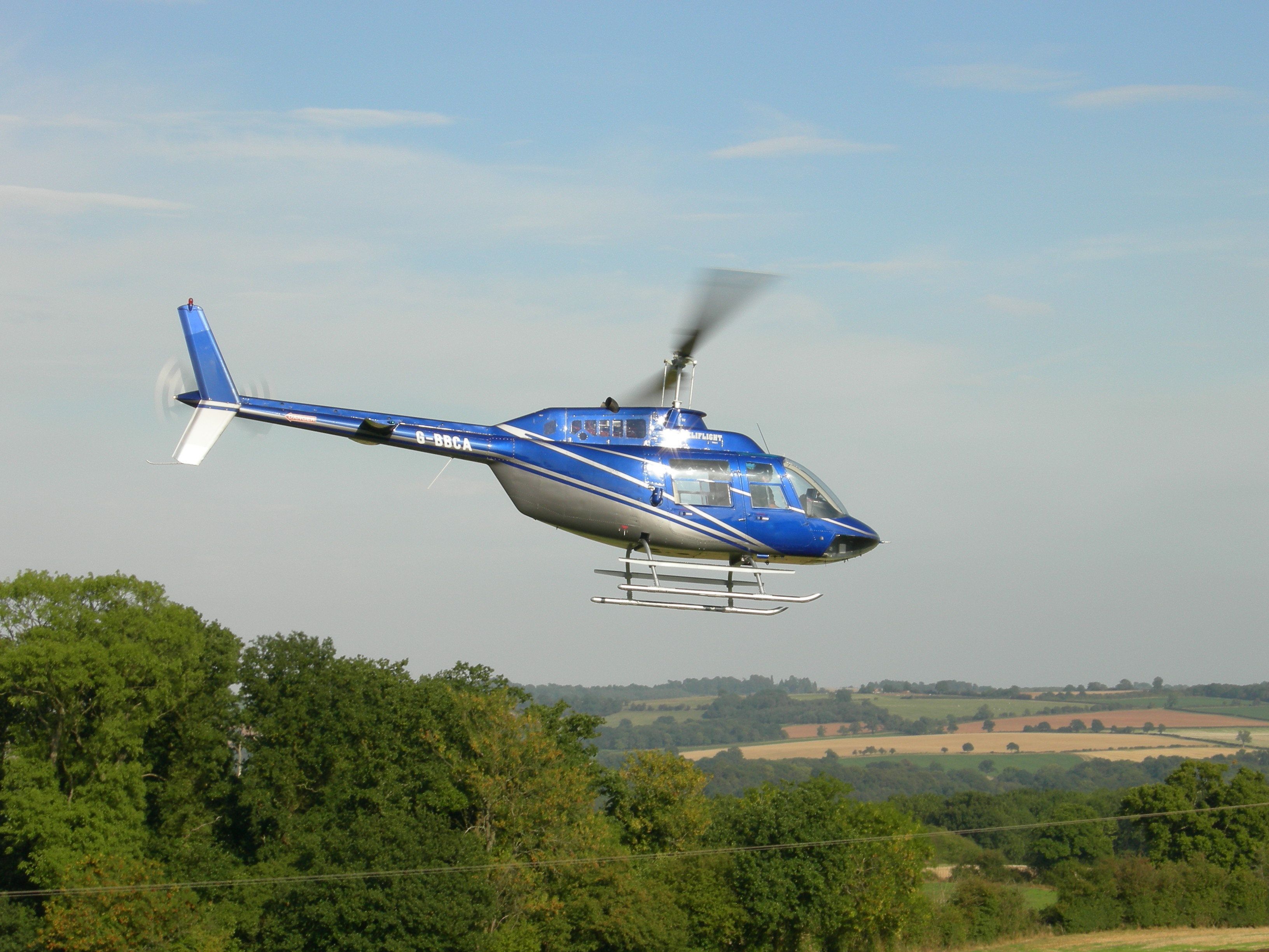 A blue helicopter with registration number G-BBCA in flight over a landscape with trees and fields.